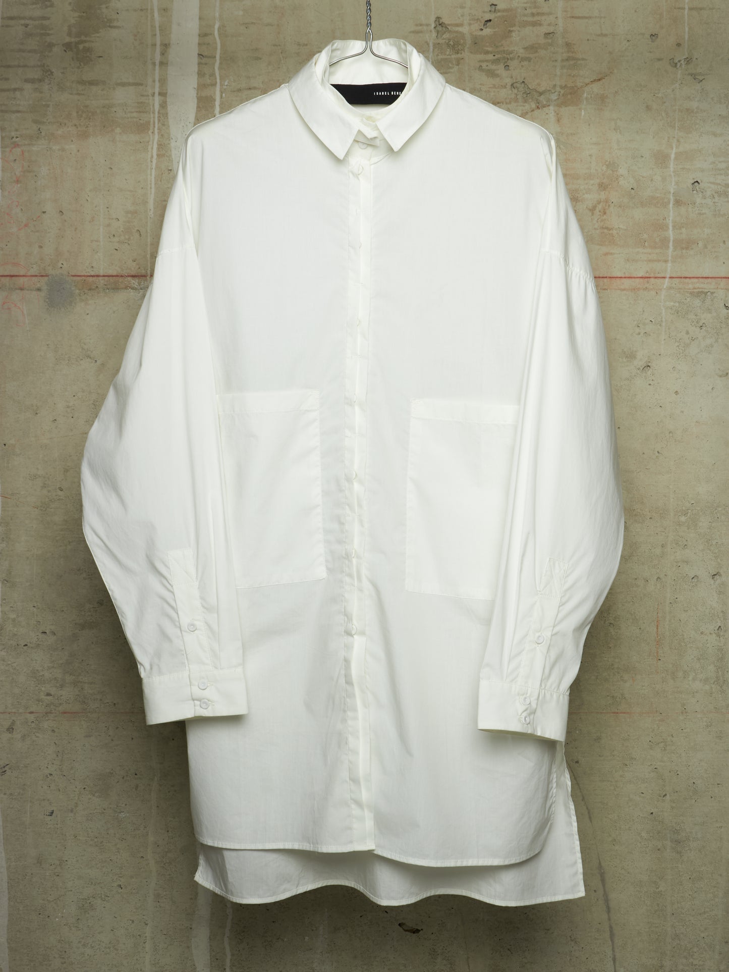 A white oversized long-sleeve button-up shirt with large front patch pockets, hanging on a hanger against a textured concrete wall.