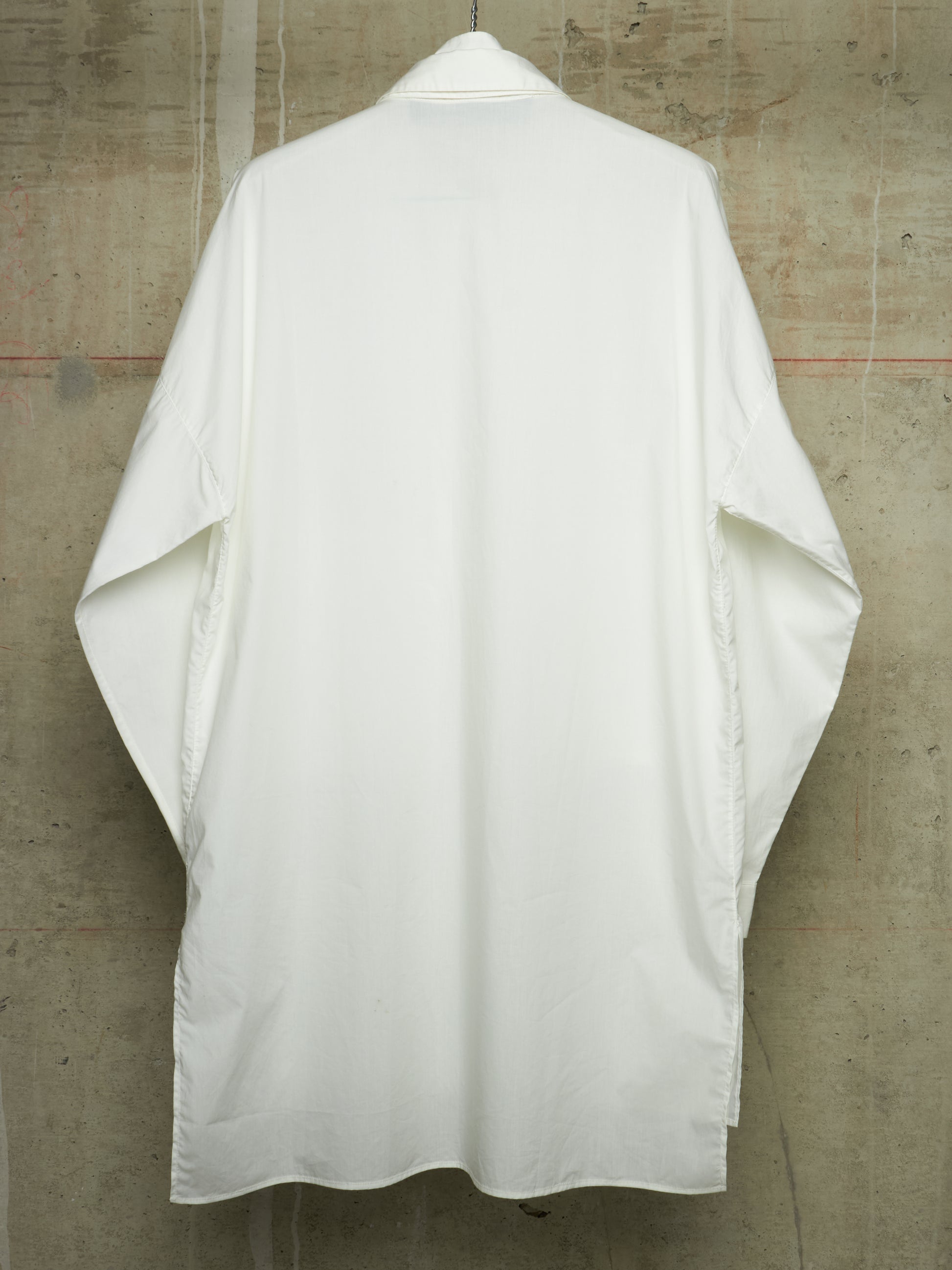 A white kimono-style shirt with wide sleeves, shown from the back hanging against a textured concrete wall.