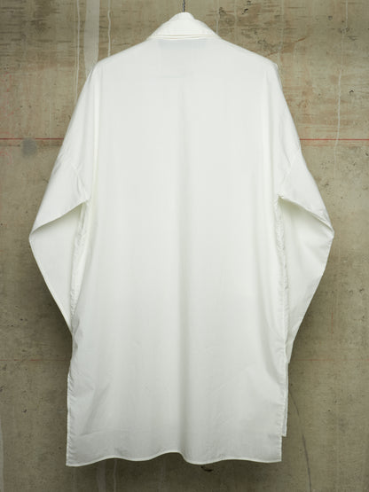 A white kimono-style shirt with wide sleeves, shown from the back hanging against a textured concrete wall.