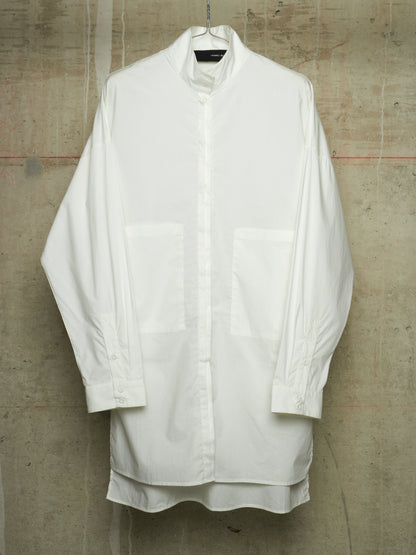 A white oversized long-sleeve button-up shirt with a high collar and two large front patch pockets, hanging against a textured concrete wall.
