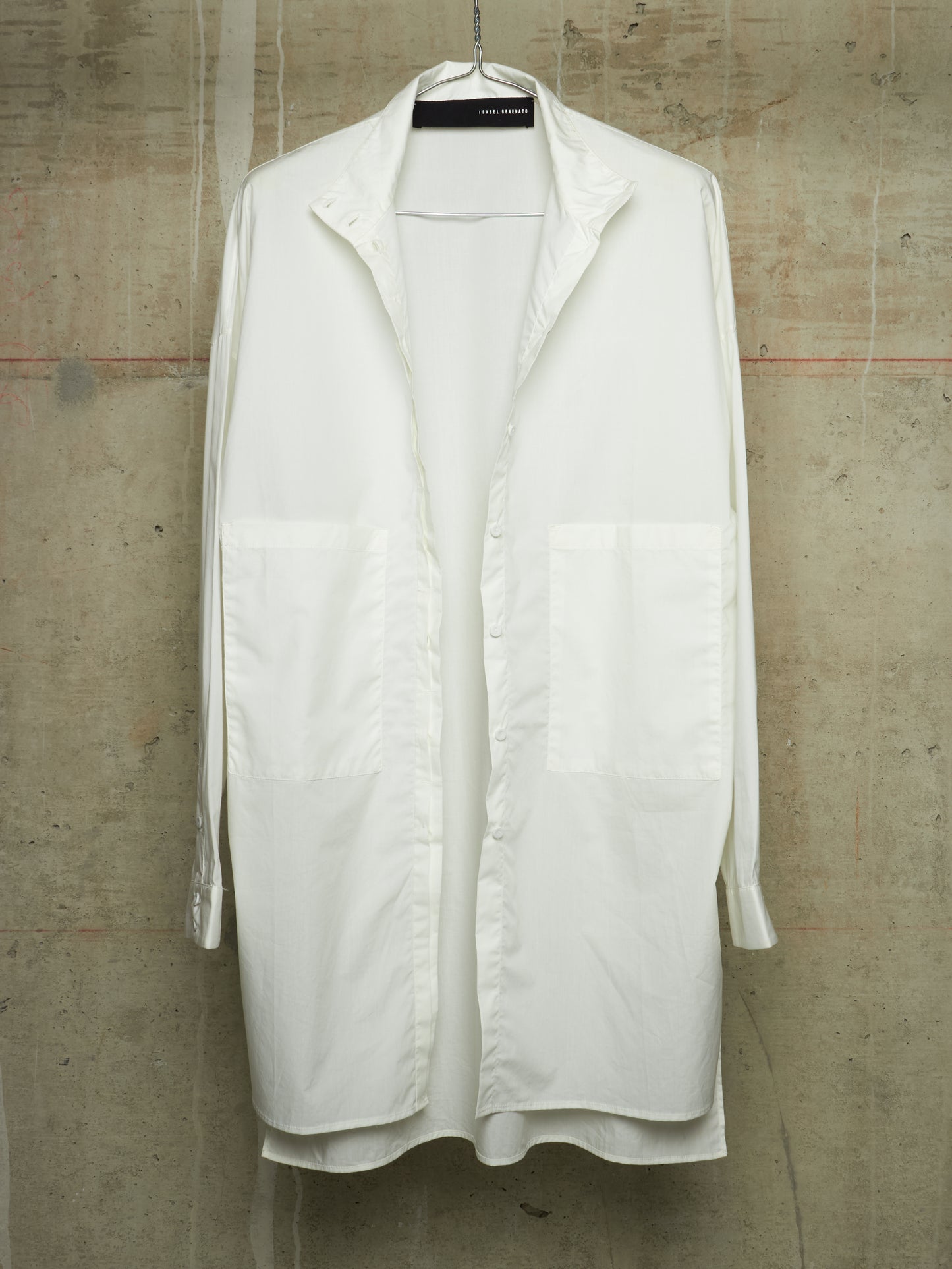 A white long-sleeve button-up shirt with large front patch pockets, hanging on a black hanger against a textured concrete wall.