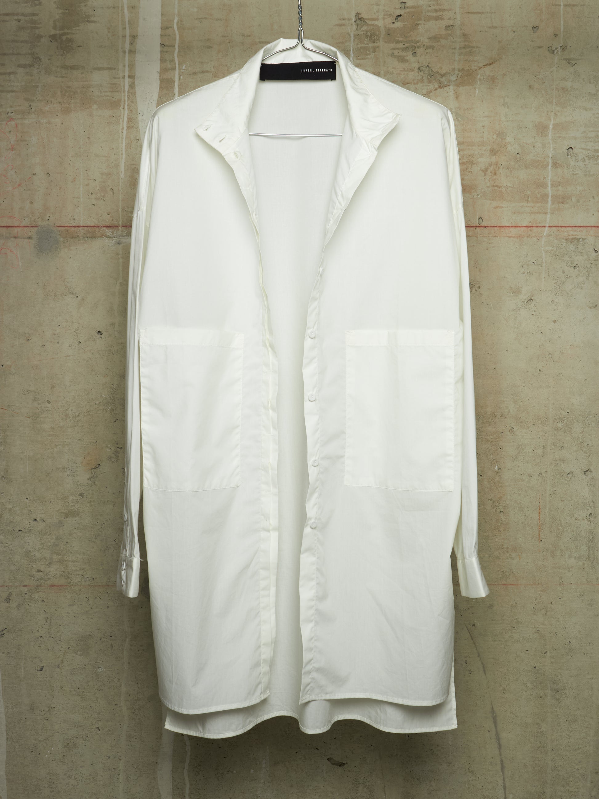A white long-sleeve button-up shirt with large front patch pockets, hanging on a black hanger against a textured concrete wall.