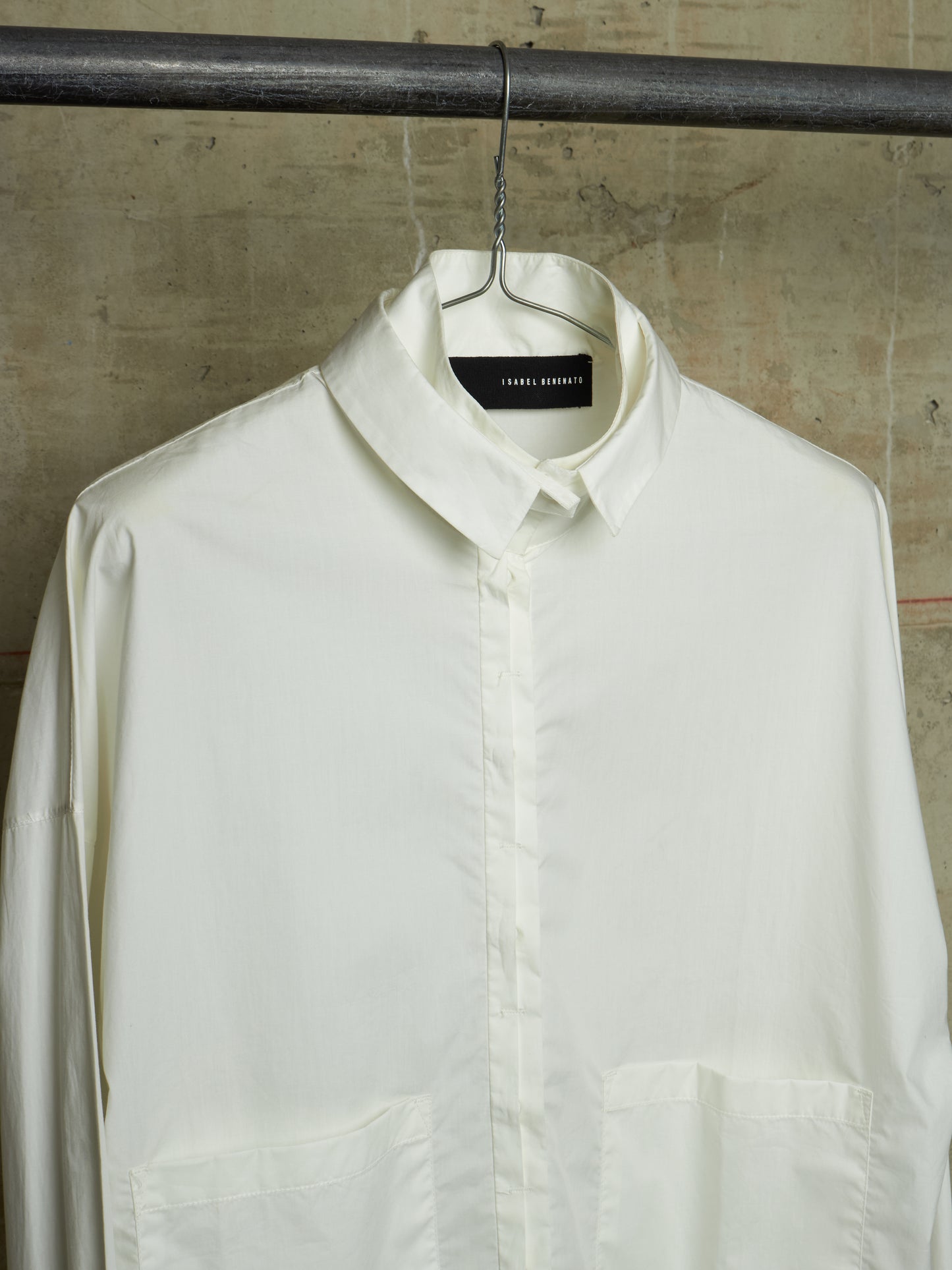 An off-white long-sleeve button-down shirt by Tommy Hilfiger, hanging on a black metal hanger from a dark industrial rod against a textured concrete wall.