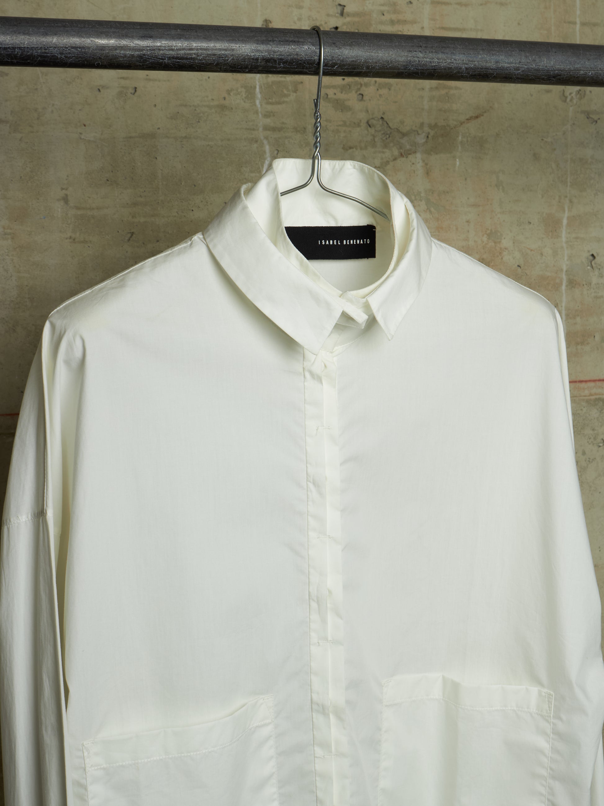 An off-white long-sleeve button-down shirt by Tommy Hilfiger, hanging on a black metal hanger from a dark industrial rod against a textured concrete wall.
