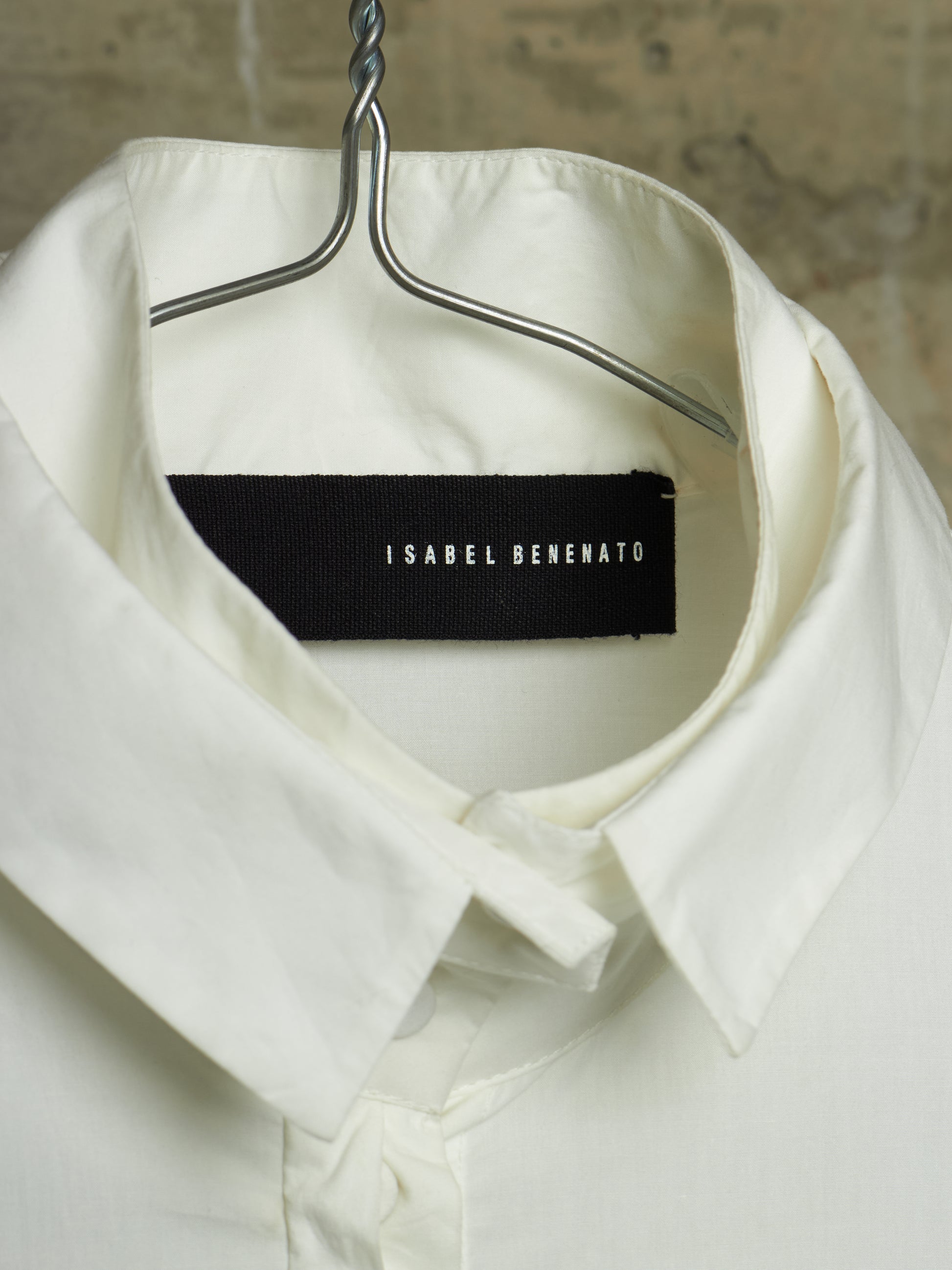 An off-white collared shirt hanging on a metal hanger against a textured stone wall background. The inside collar features a black fabric brand label with the text 'ISABEL BENENATO' in white.