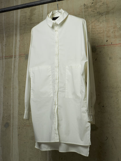 A long-sleeve white button-up shirt dress with large front patch pockets, hanging on a metal hanger against a textured concrete wall.