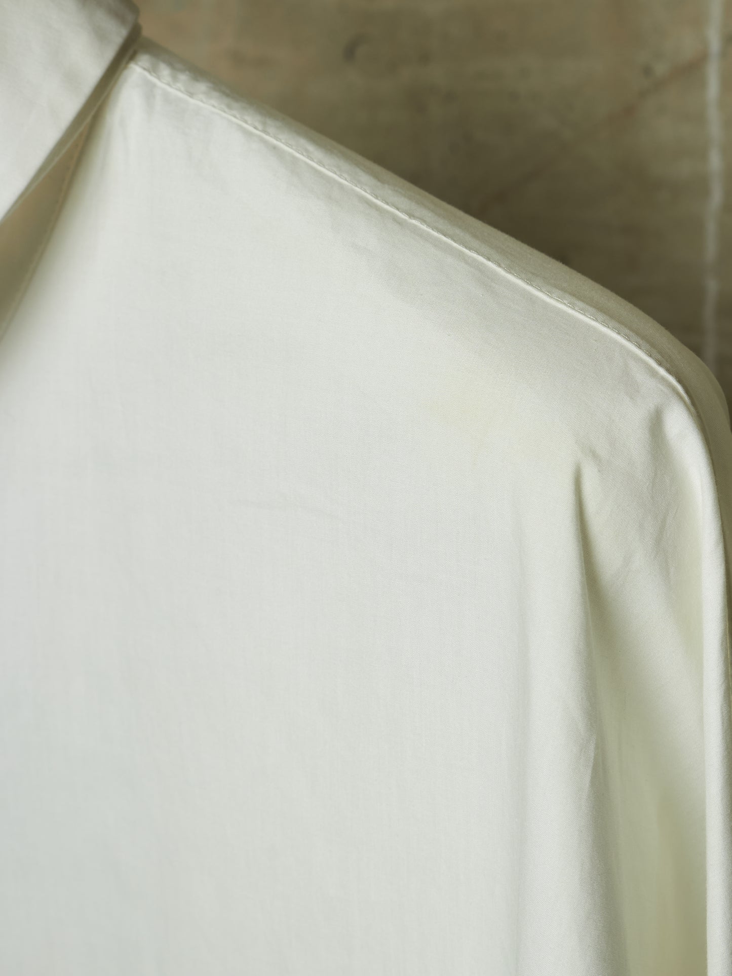 A close-up of a white or off-white fabric garment, showing the collar and shoulder area with soft folds and a neutral concrete-like background.
