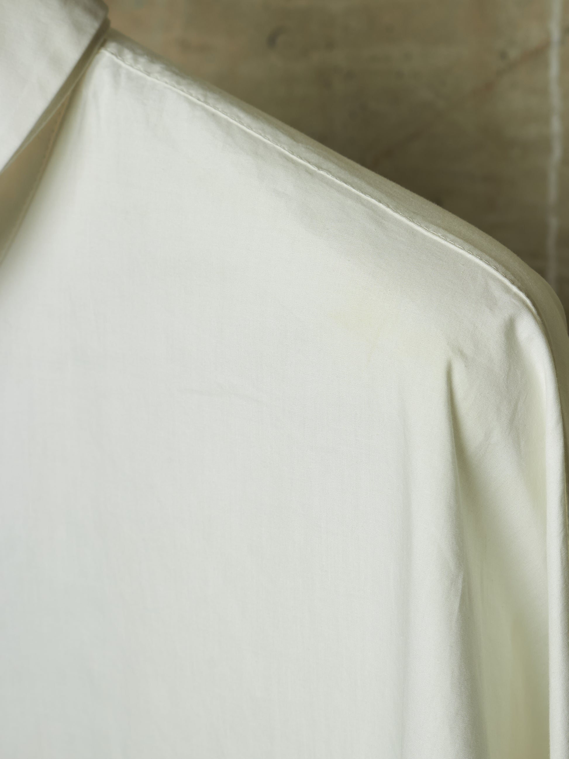 A close-up of a white or off-white fabric garment, showing the collar and shoulder area with soft folds and a neutral concrete-like background.