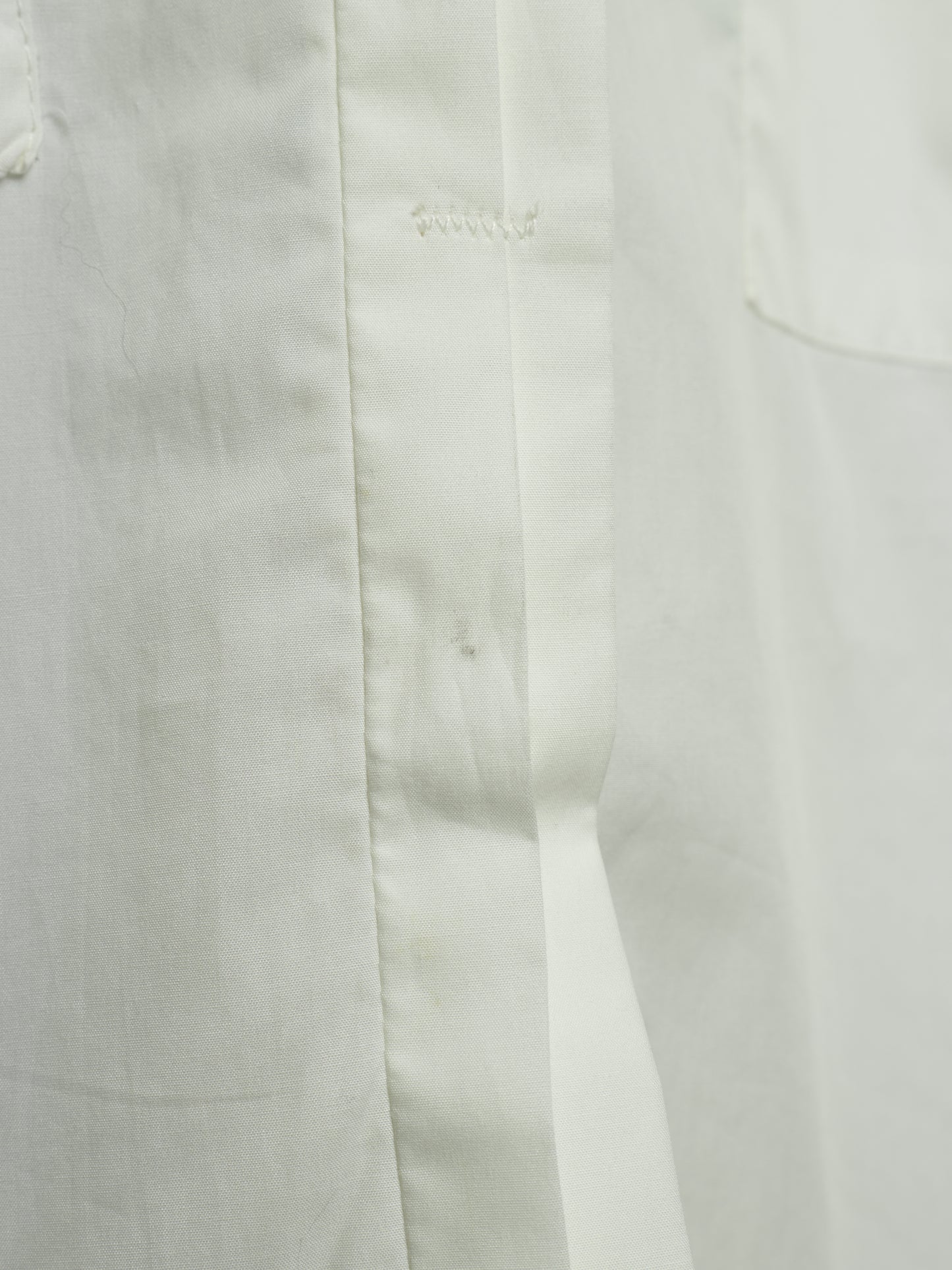 Close-up detail of a white fabric garment showing a vertical seam, button placket stitching, and a pocket edge.