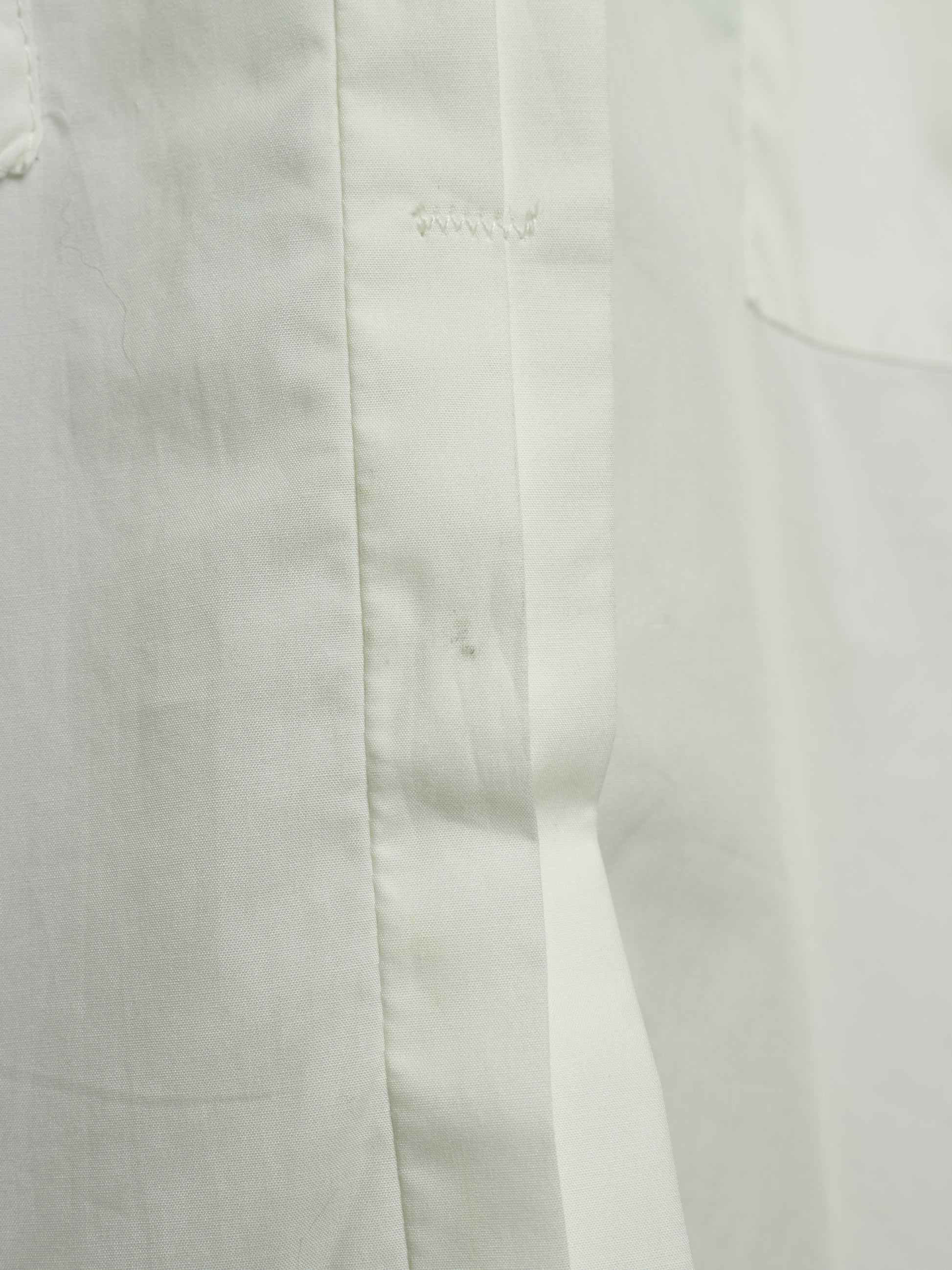 Close-up detail of a white fabric garment showing a vertical seam, button placket stitching, and a pocket edge.
