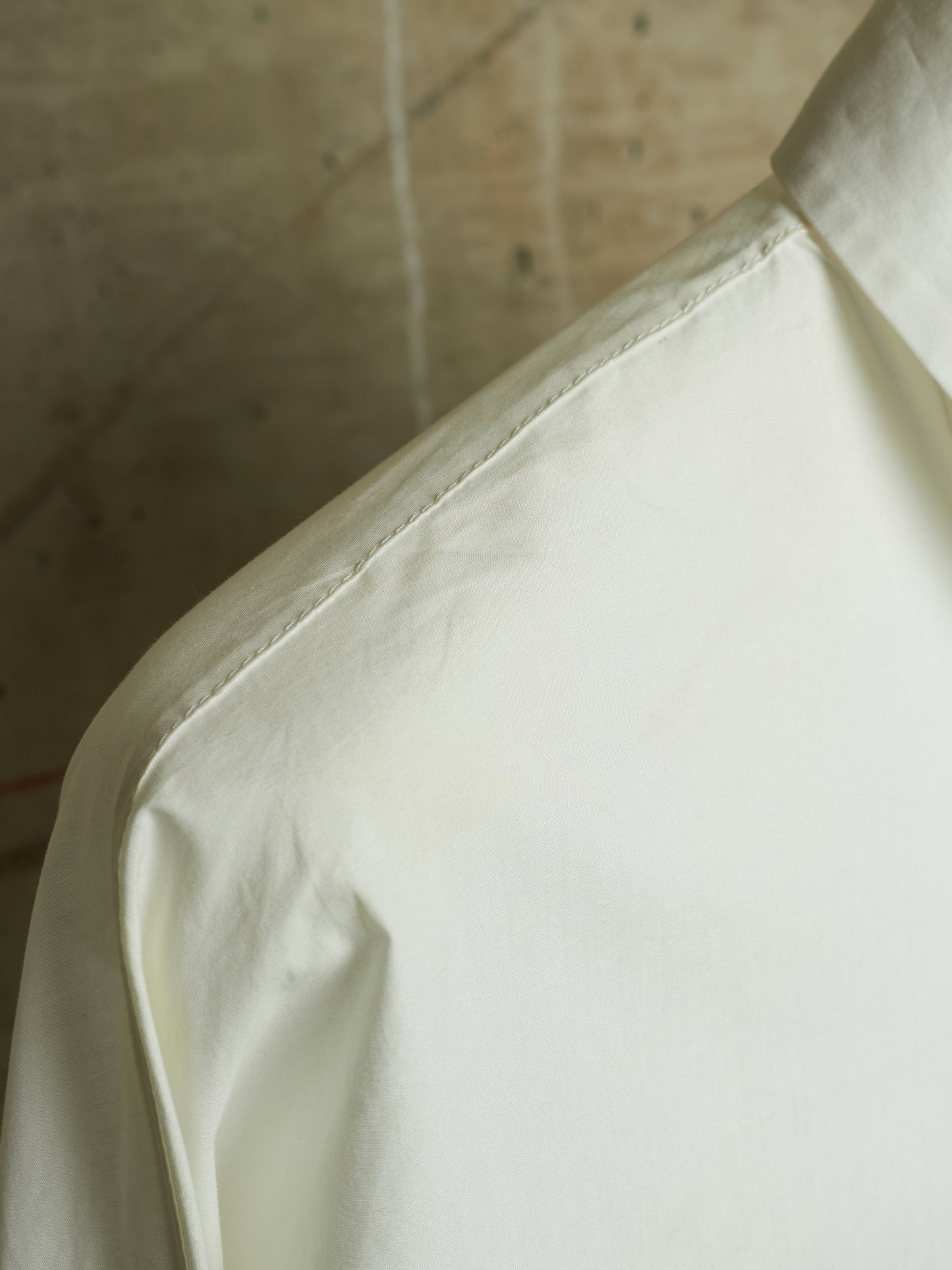 Close-up of a light cream or off-white fabric garment, showing the shoulder seam and collar area against a neutral textured background.