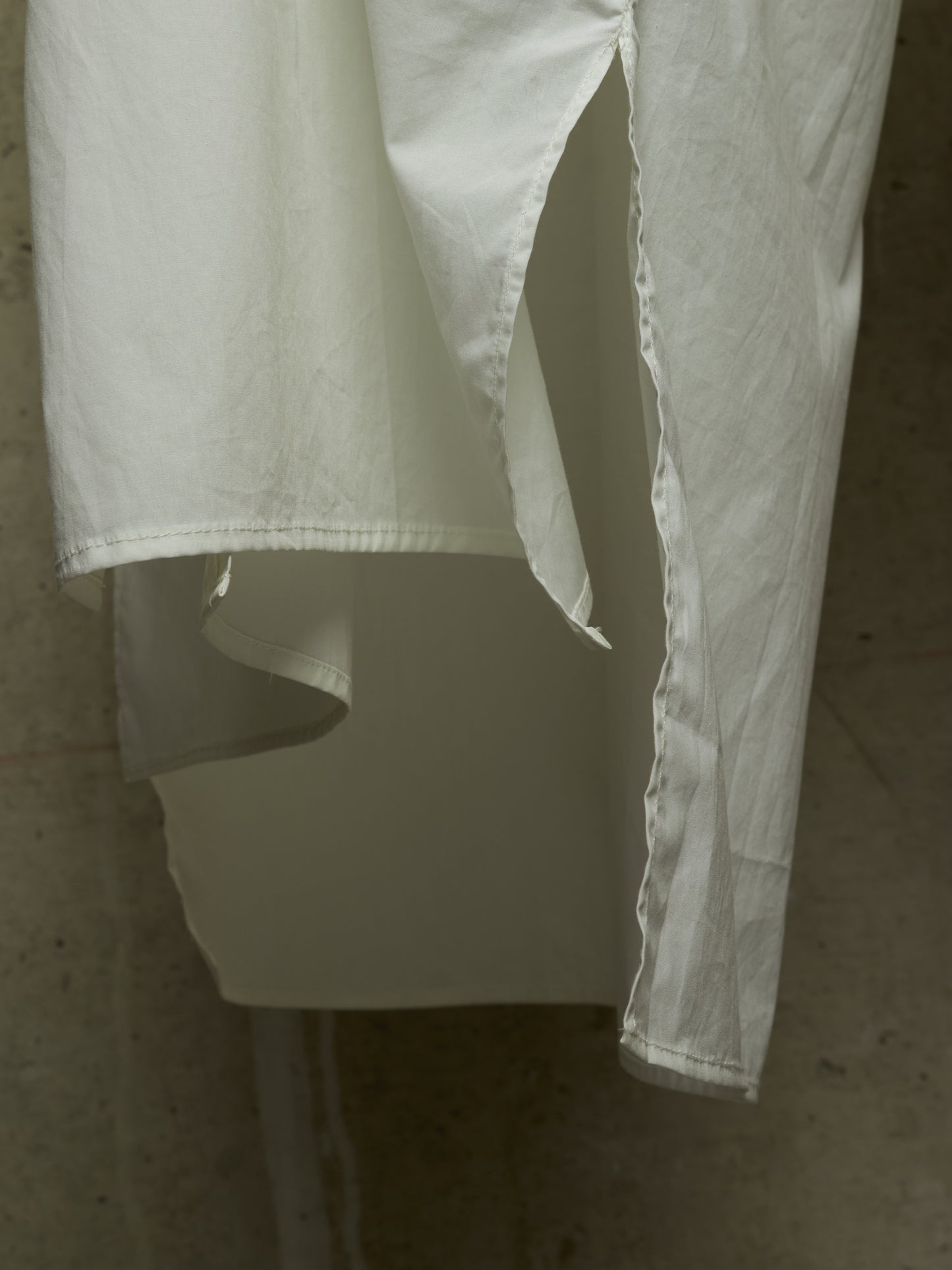 A close-up of a white or off-white garment hanging against a textured beige wall, showing the fabric drape, hem, and side slit detail.