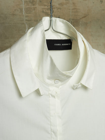 A close-up of a white collared button-up shirt hanging on a metal hanger against a textured beige wall. The neck label reads ISABEL RONDONO.