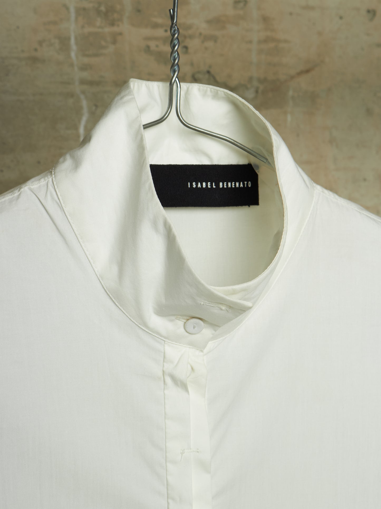 A close-up of a white collared shirt hanging on a metal hanger against a textured beige wall. The brand label 'ISABEL MARANT' is visible inside the collar.