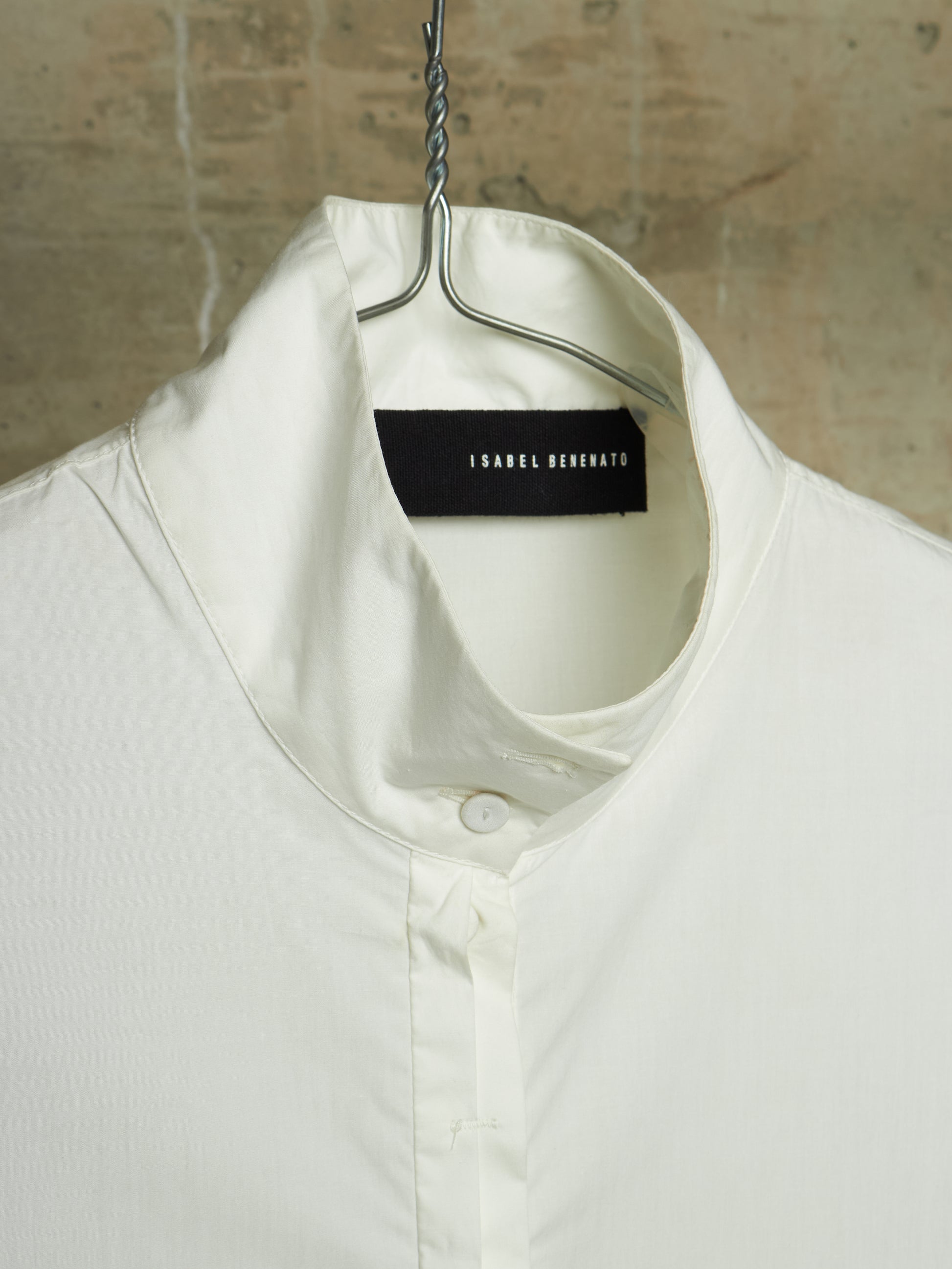 A close-up of a white collared shirt hanging on a metal hanger against a textured beige wall. The brand label 'ISABEL MARANT' is visible inside the collar.
