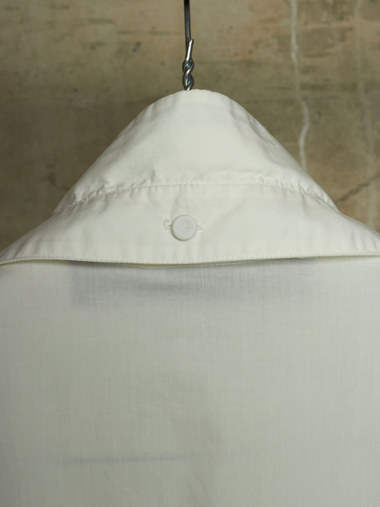 Close-up of the collar of an off-white or cream-colored shirt hanging on a metal hanger against a textured beige wall.