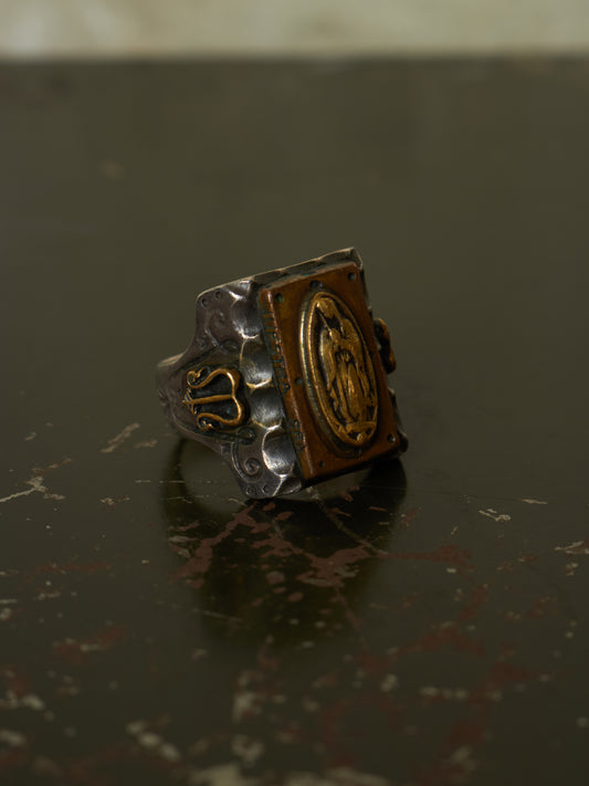 A square signet ring with a silver-toned textured band and gold-toned detailing, including an oval medallion on the face, resting on a dark reflective surface.
