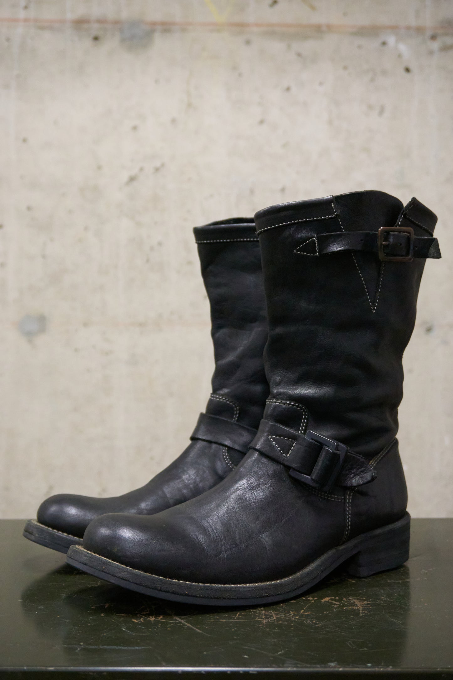 Pair of black leather boots with buckles on a reflective surface and concrete wall background