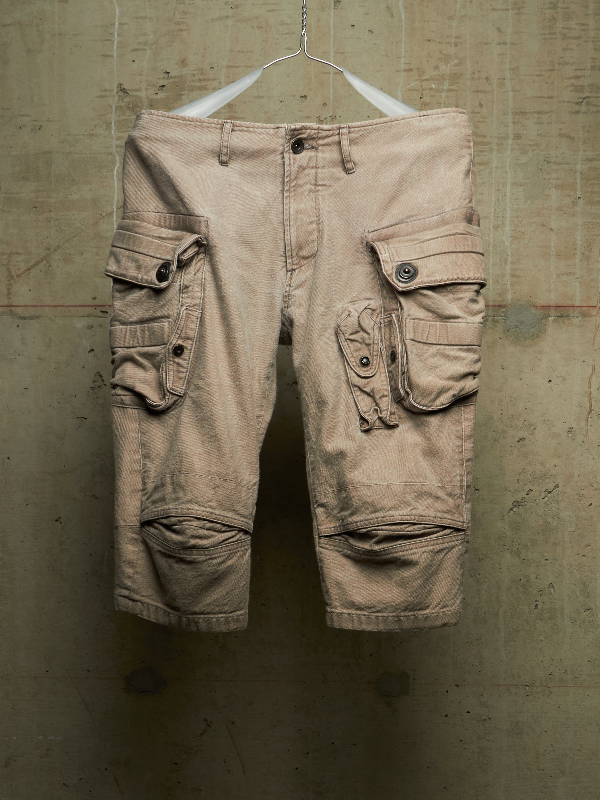 A pair of beige cargo shorts with multiple flap pockets and knee patches, hanging from a white hanger against a textured concrete wall.