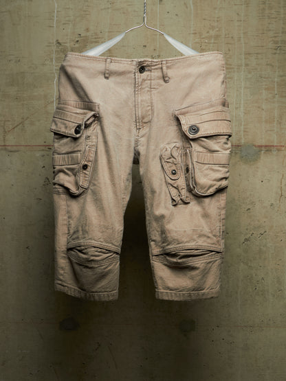 A pair of beige cargo shorts with multiple flap pockets and knee patches, hanging from a white hanger against a textured concrete wall.