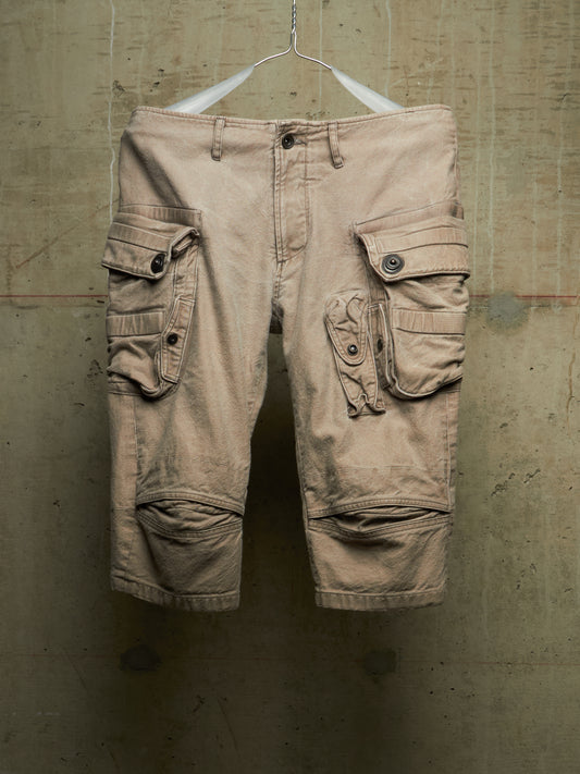 A pair of beige cargo shorts with multiple flap pockets and knee patches, hanging from a white hanger against a textured concrete wall.