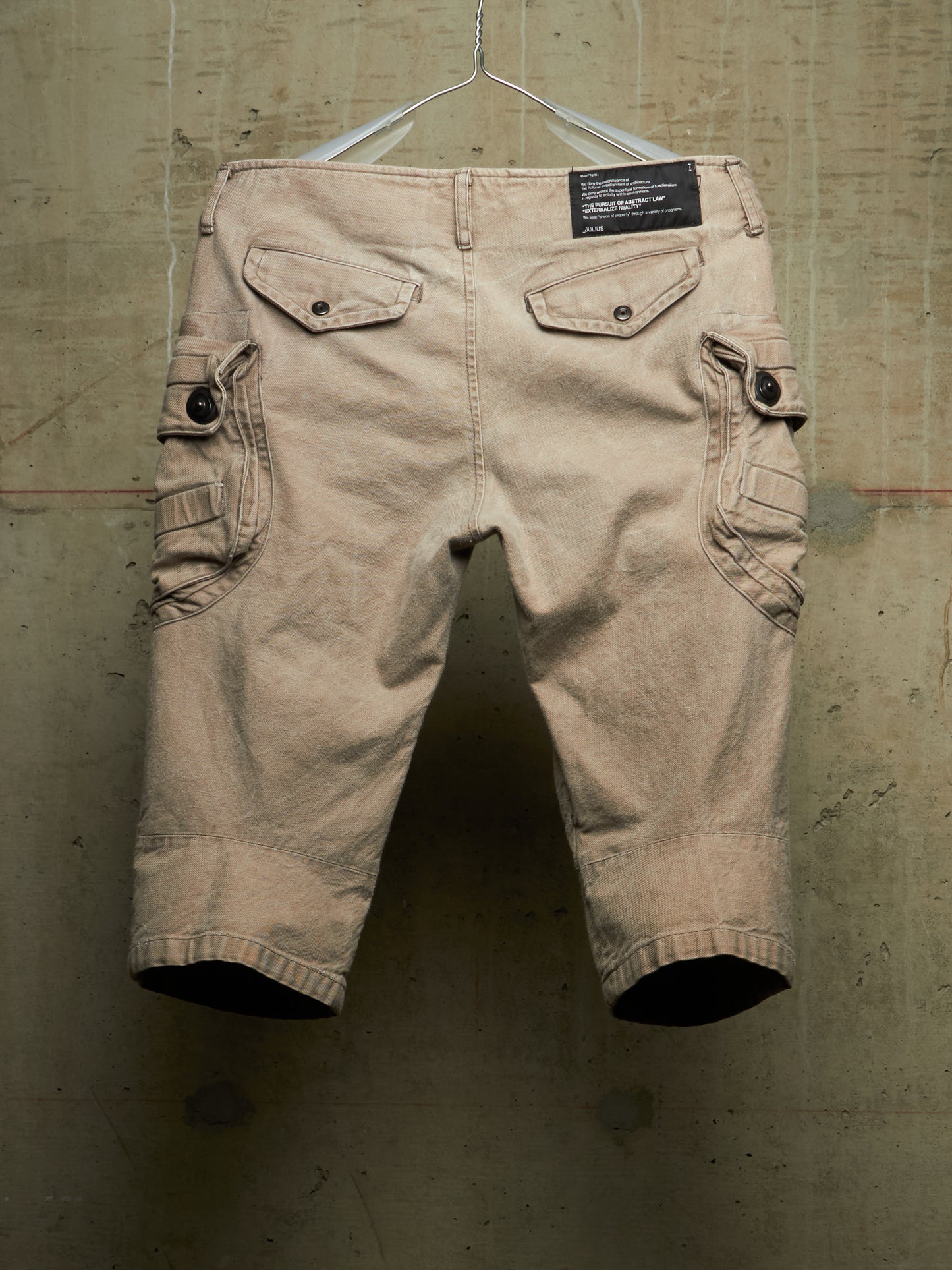 A pair of beige cargo shorts with flap pockets and button details, hanging from a metal hanger against a textured concrete wall.