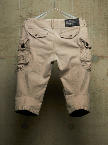 A pair of beige cargo shorts with flap pockets and button details, hanging from a metal hanger against a textured concrete wall.