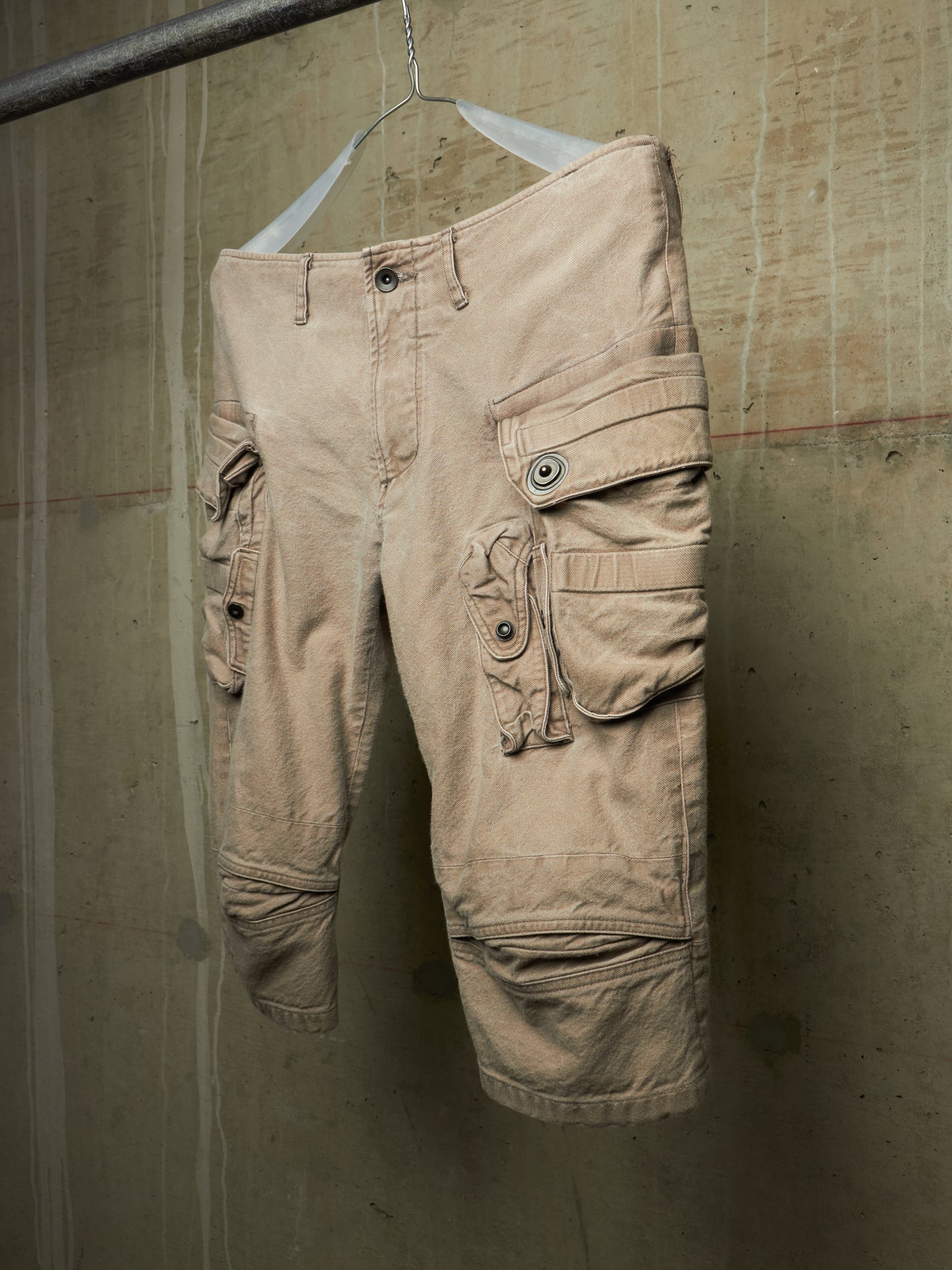 A pair of beige cargo shorts with multiple flap pockets and button details, hanging on a white hanger against a textured concrete wall.