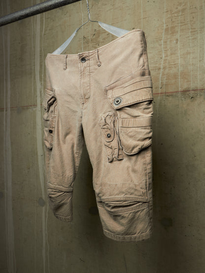 A pair of beige cargo shorts with multiple flap pockets and button details, hanging on a white hanger against a textured concrete wall.