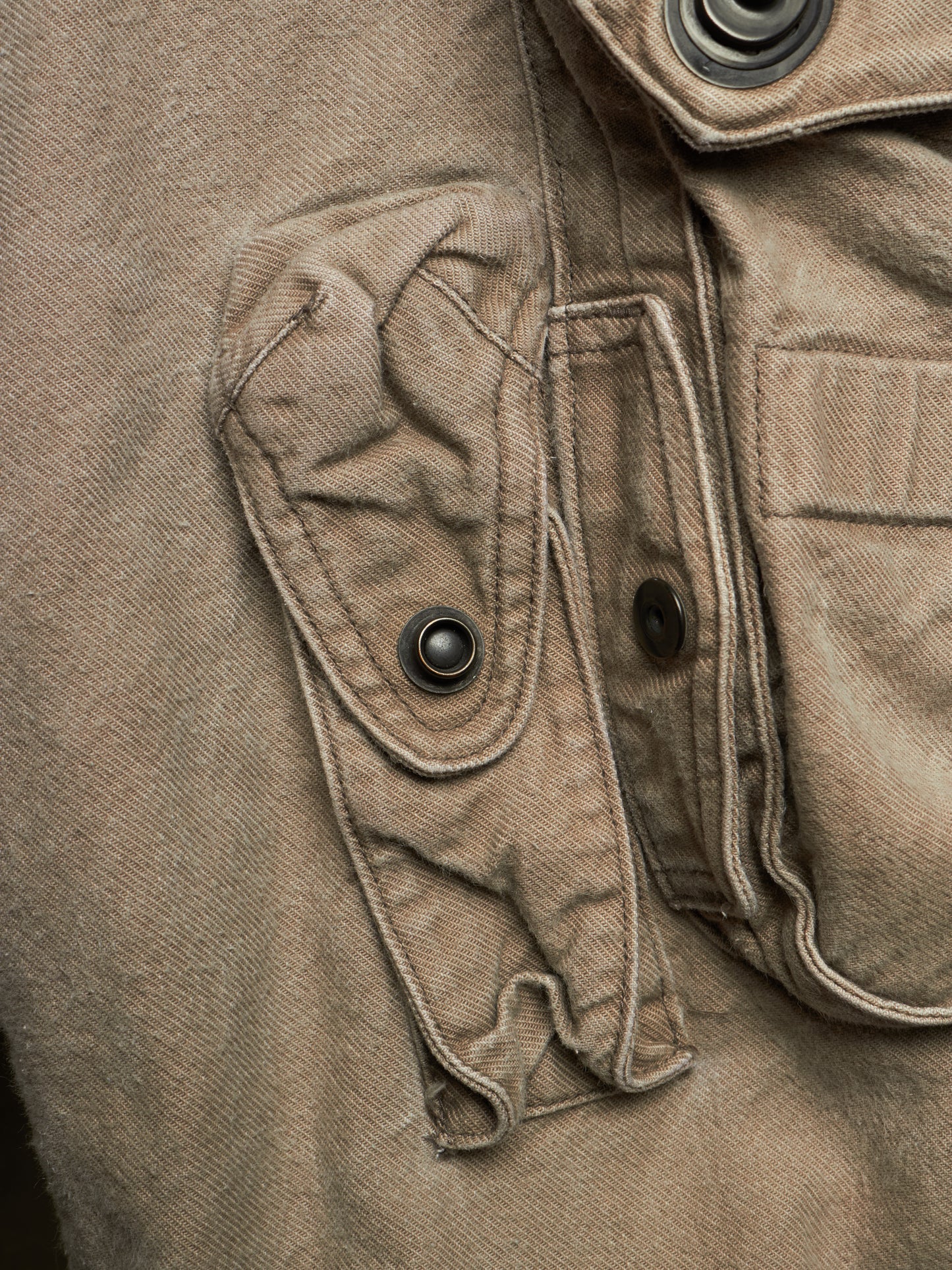 Close-up detail of a brown canvas utility jacket showing a buttoned flap pocket and metal snap buttons.