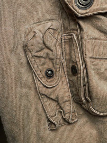 Close-up detail of a brown canvas utility jacket showing a buttoned flap pocket and metal snap buttons.