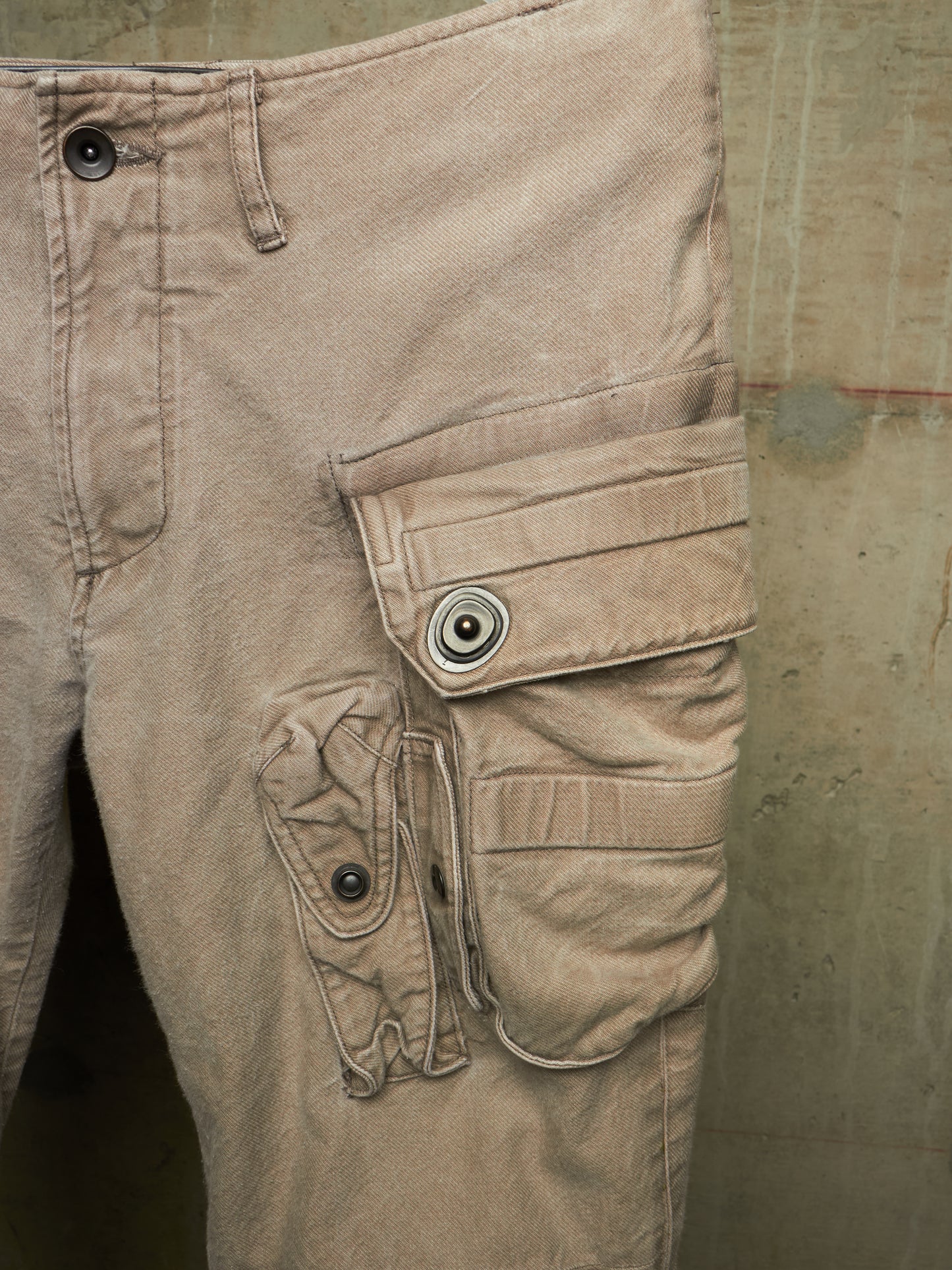 Close-up of tan cargo pants showing the right leg with multiple flap cargo pockets and metal snap buttons, photographed against a textured concrete wall.