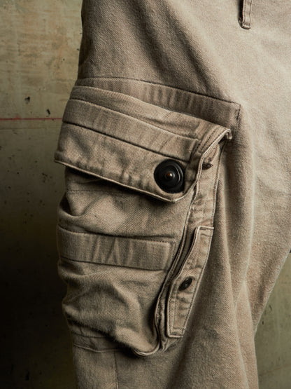 Close-up of a beige cargo pant leg showing a large buttoned flap pocket and a buttoned side pocket against a textured concrete wall.