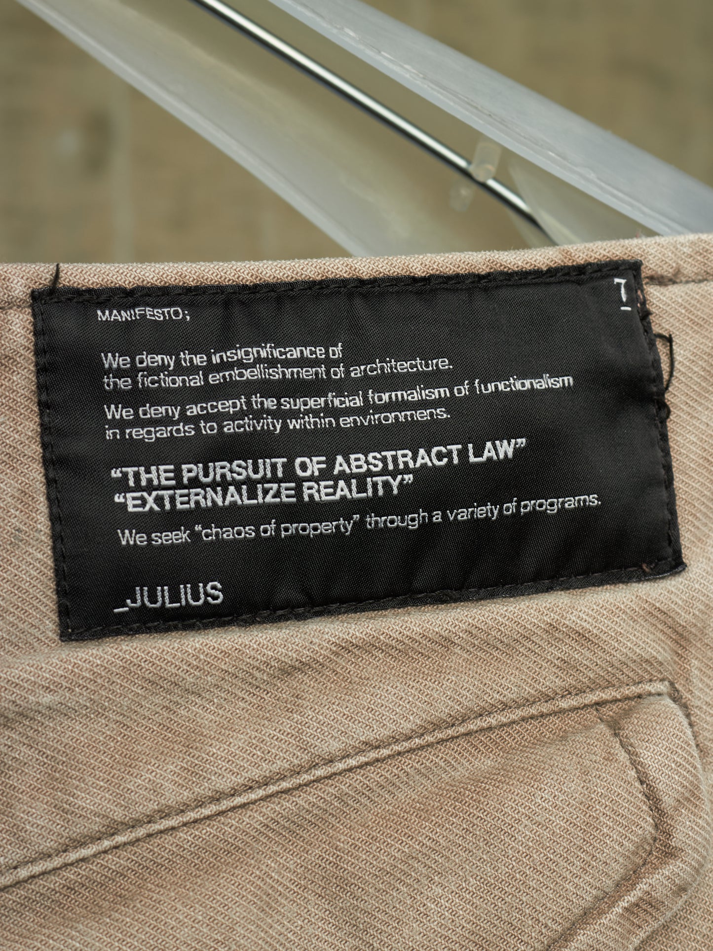 Close-up of a beige woven textile item with a black sewn label reading JULIUS and philosophical text, photographed against a softly blurred gray background.