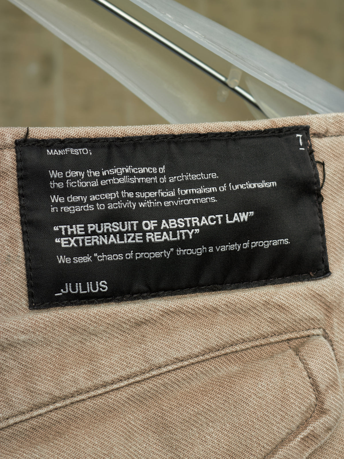 Close-up of a beige woven textile item with a black sewn label reading JULIUS and philosophical text, photographed against a softly blurred gray background.