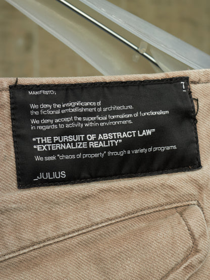 Close-up of a beige woven textile item with a black sewn label reading JULIUS and philosophical text, photographed against a softly blurred gray background.