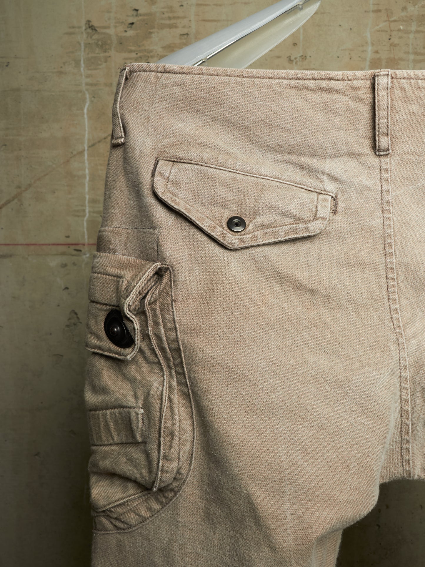 Close-up of beige cargo shorts showing the back pocket details, including a flap pocket with a metal snap and a side cargo pocket with a black button, set against a textured concrete wall.