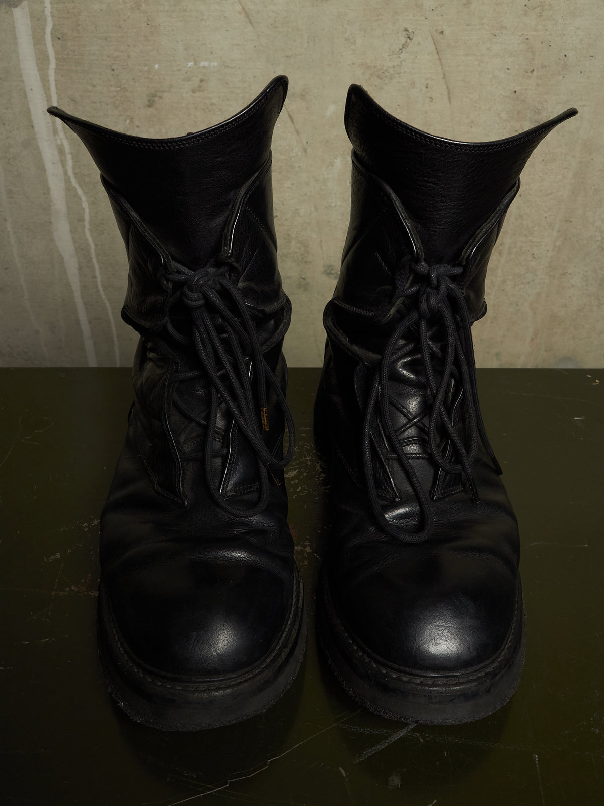 A pair of black leather lace-up combat boots with thick soles, shown from the front on a dark reflective surface against a textured beige wall.