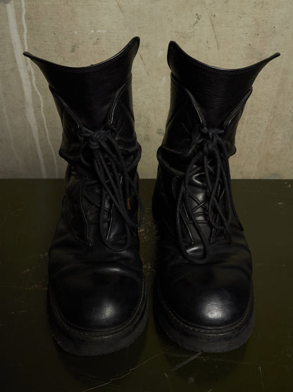 A pair of black leather lace-up combat boots with thick soles, shown from the front on a dark reflective surface against a textured beige wall.