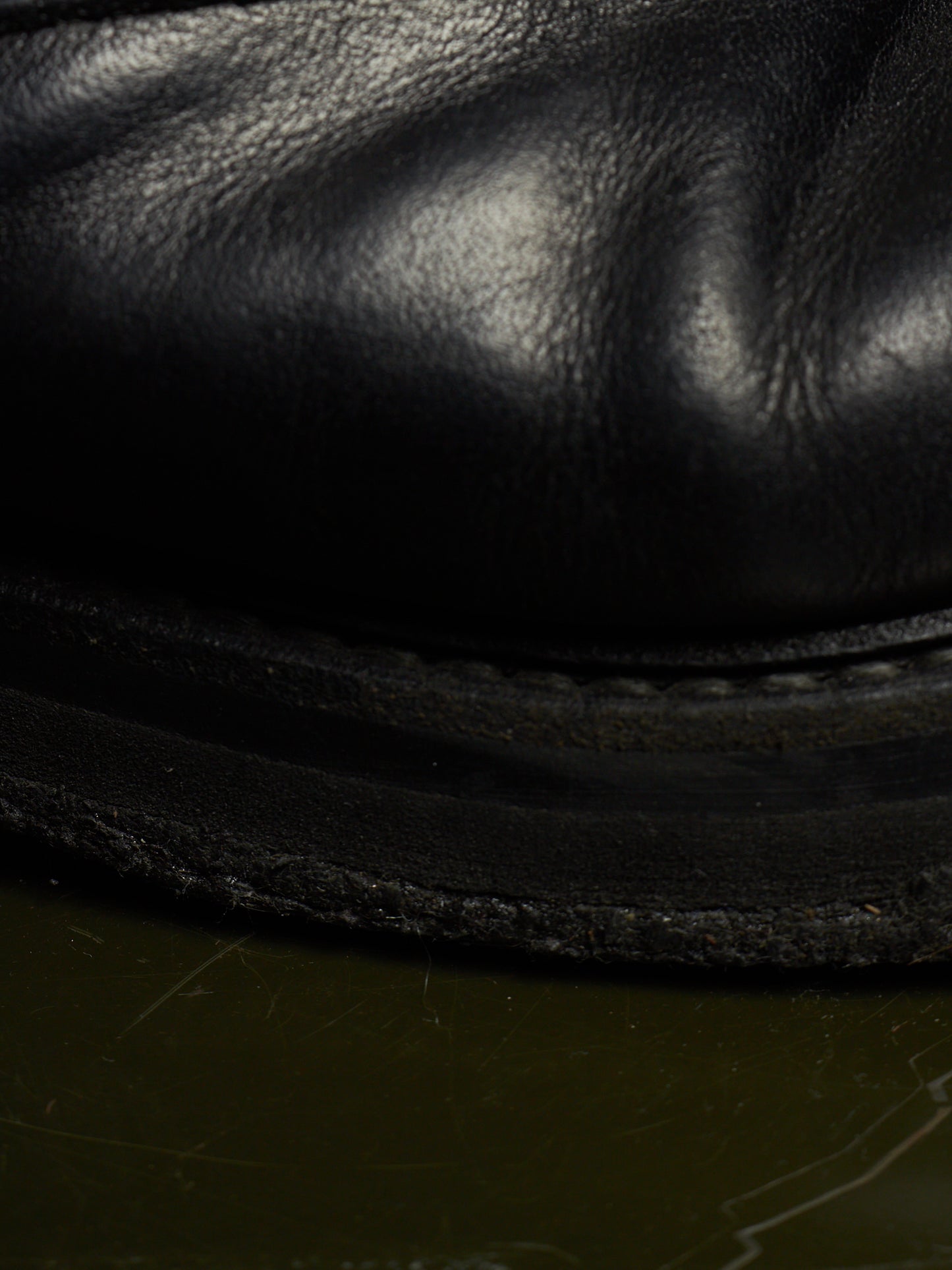 Close-up of a black leather boot or shoe sole with visible grain texture and stitching, resting on a dark olive-green surface.