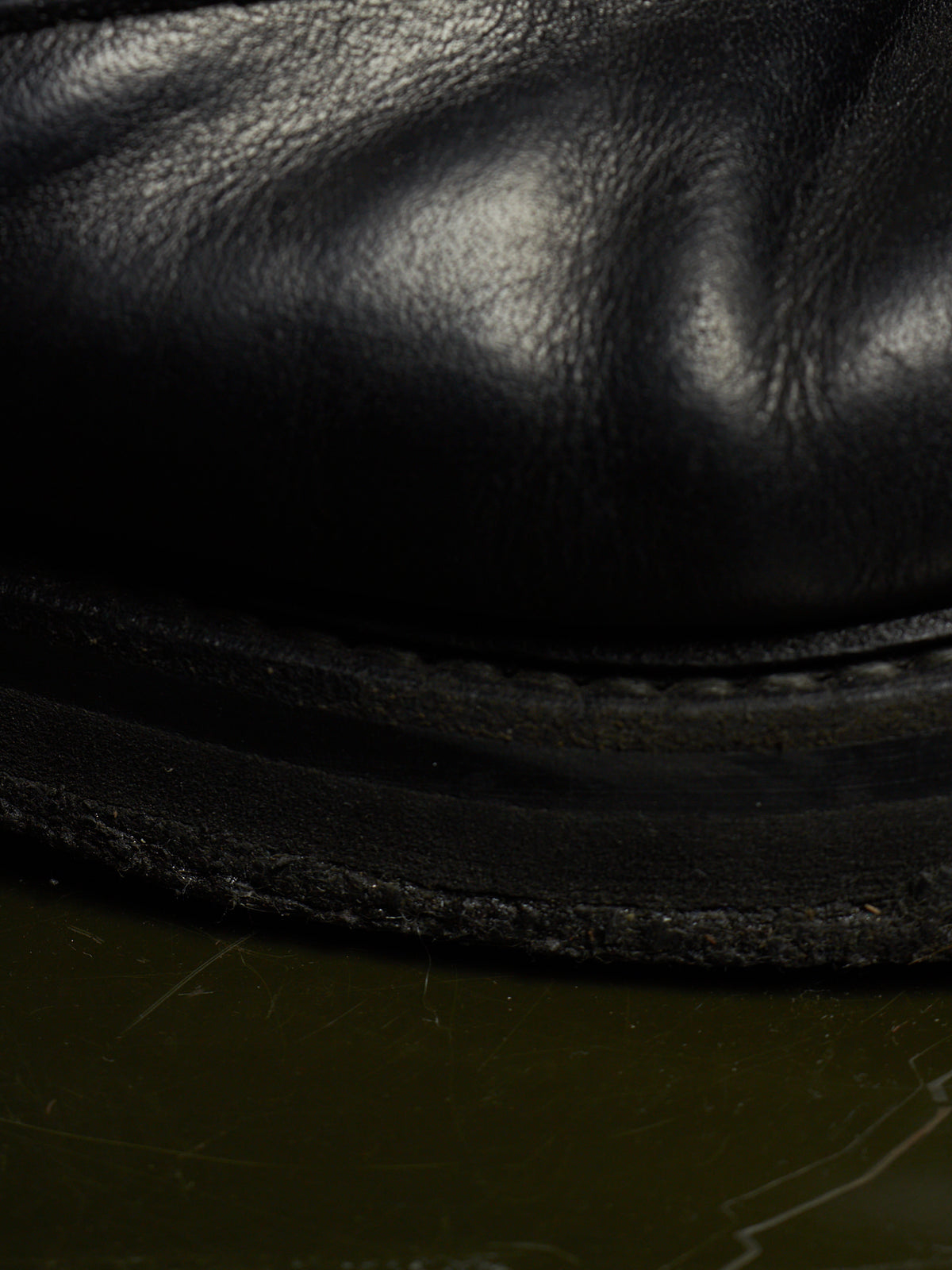 Close-up of a black leather boot or shoe sole with visible grain texture and stitching, resting on a dark olive-green surface.