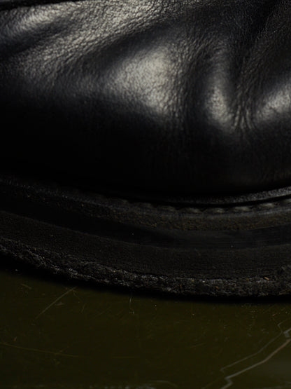 Close-up of a black leather boot or shoe sole with visible grain texture and stitching, resting on a dark olive-green surface.