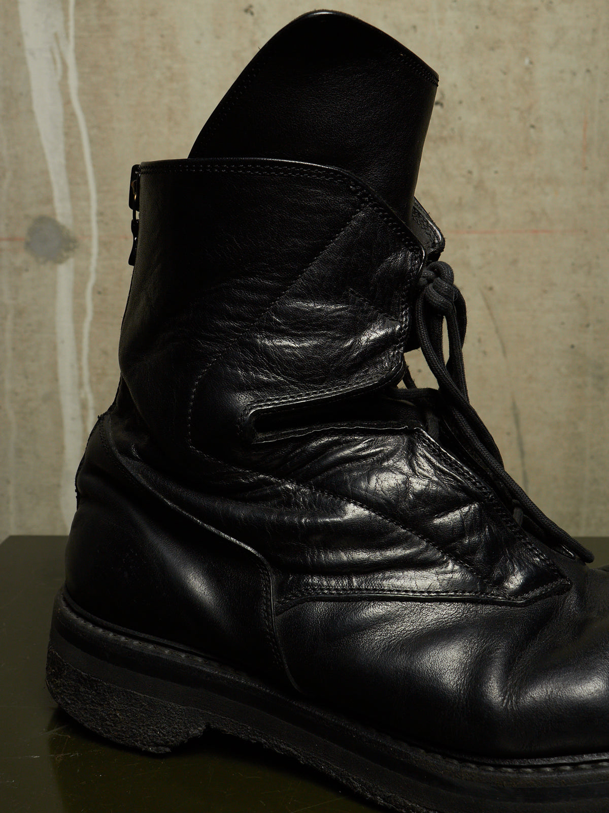 A close-up of a black leather lace-up boot with a rugged sole, set against a textured concrete wall background.