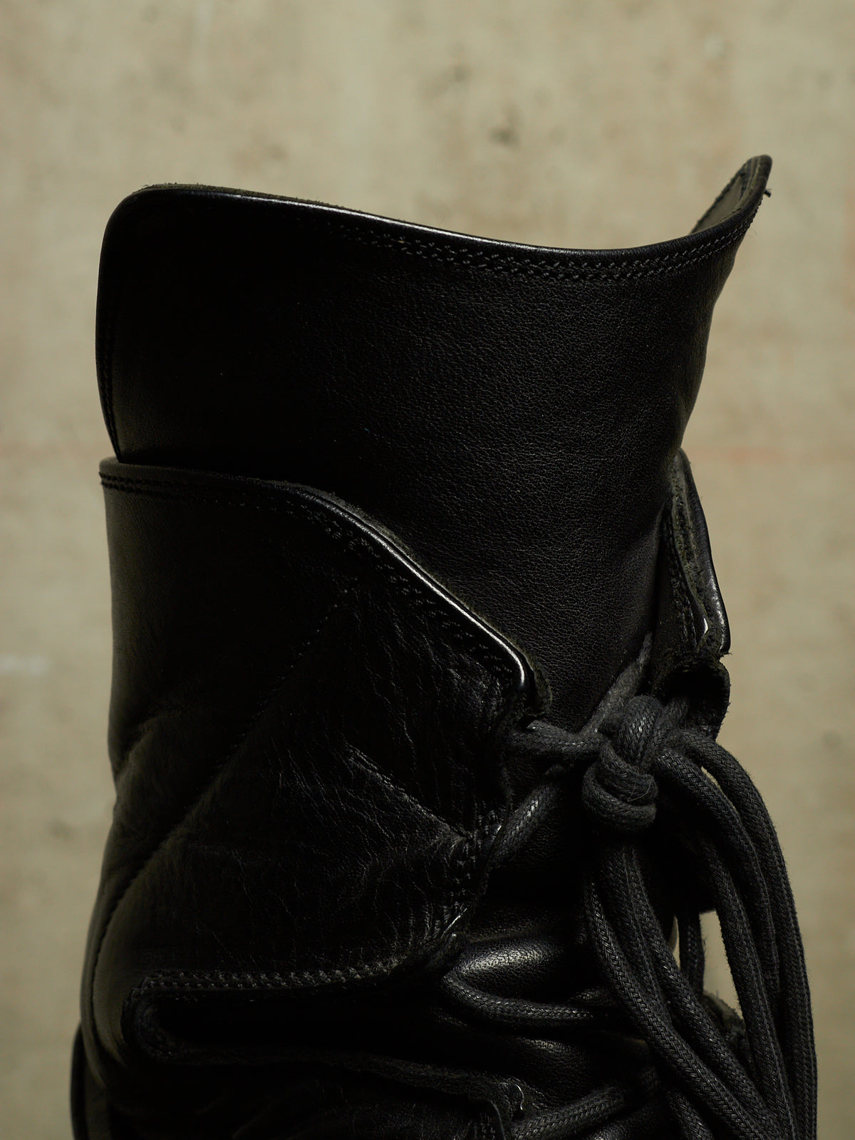 Close-up of a black leather lace-up boot against a textured beige background.