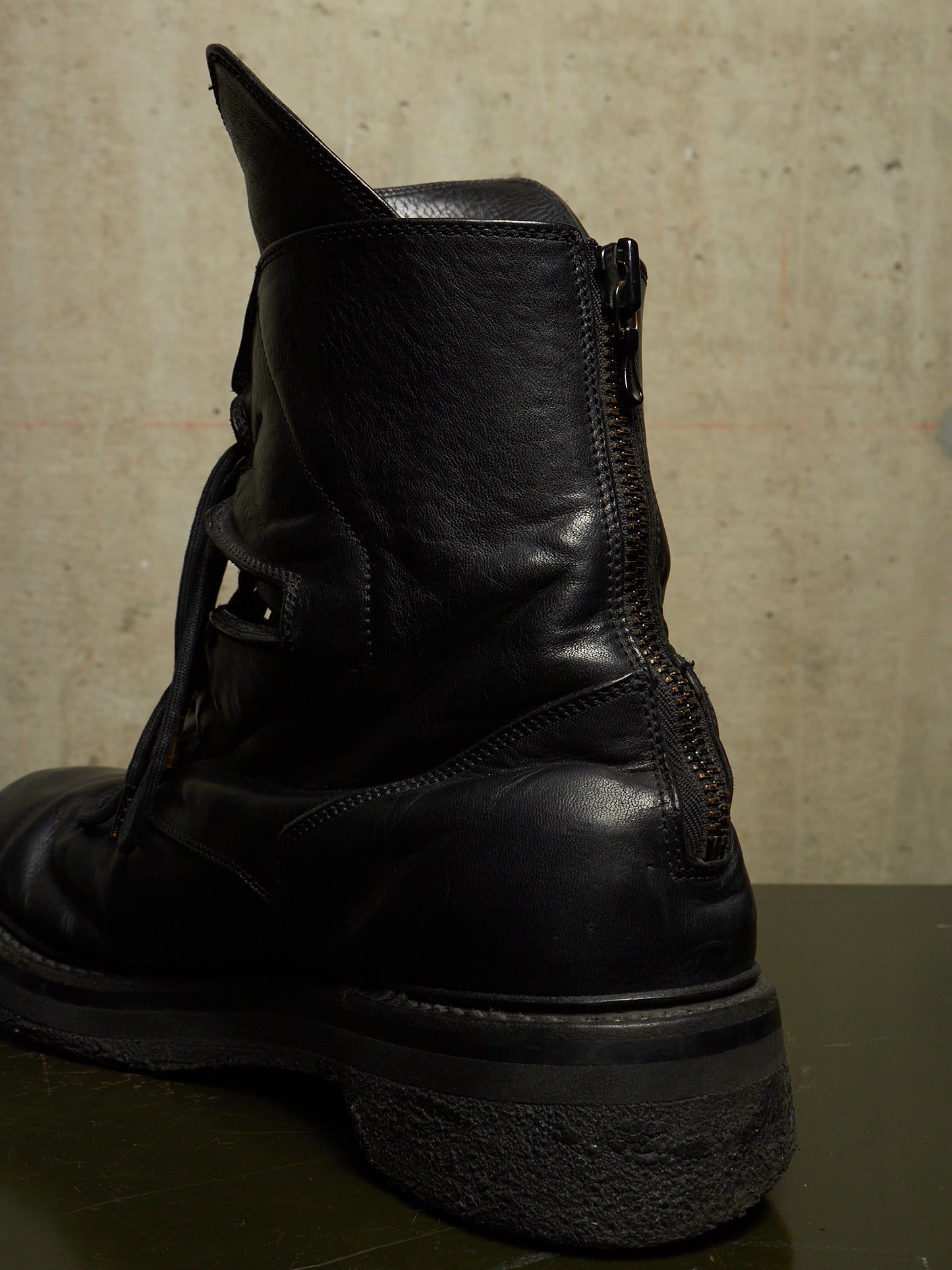 A black leather ankle boot with a side zipper, shown from a rear three-quarter angle against a textured concrete wall.