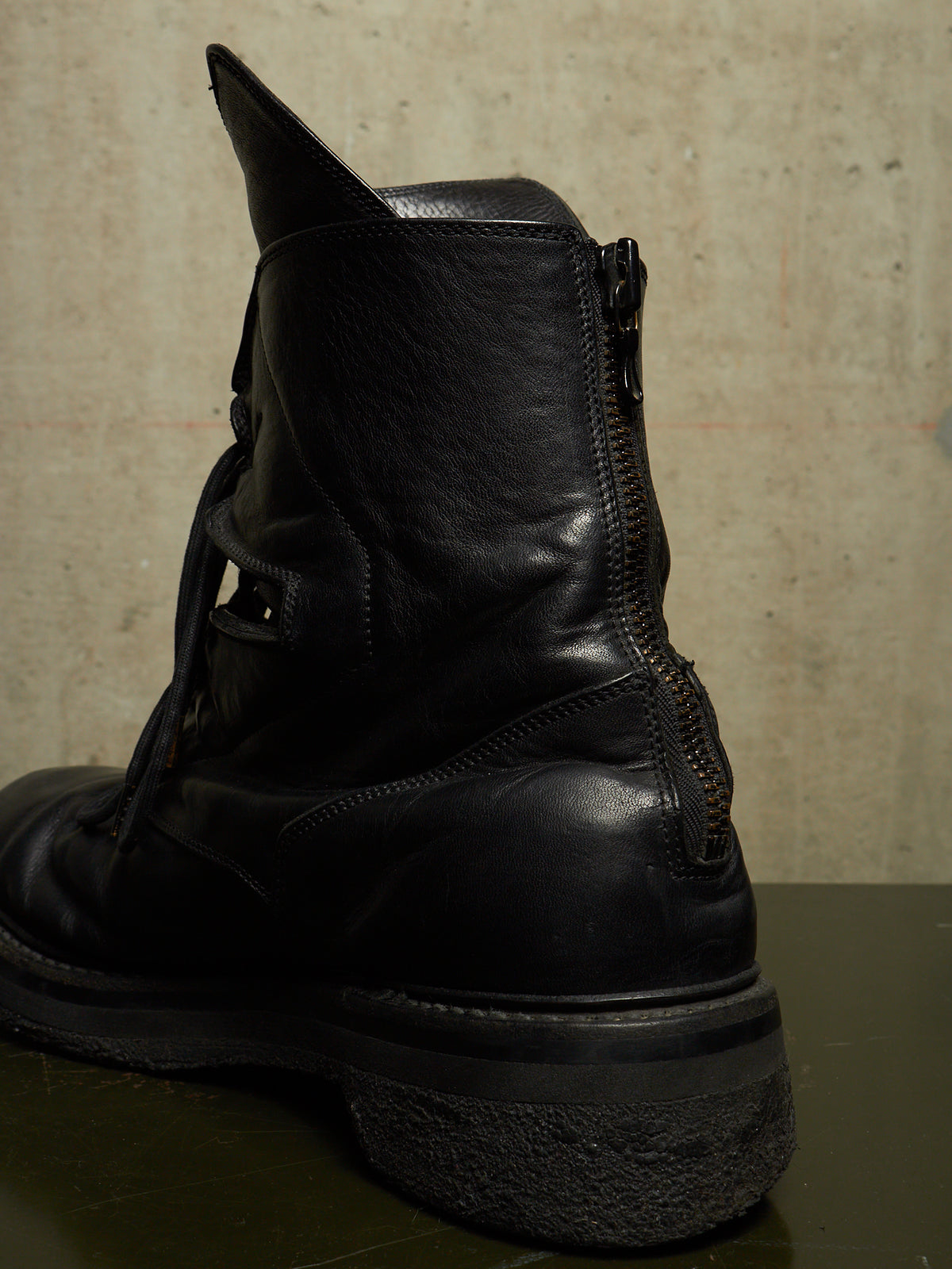 A black leather ankle boot with a side zipper, shown from a rear three-quarter angle against a textured concrete wall.