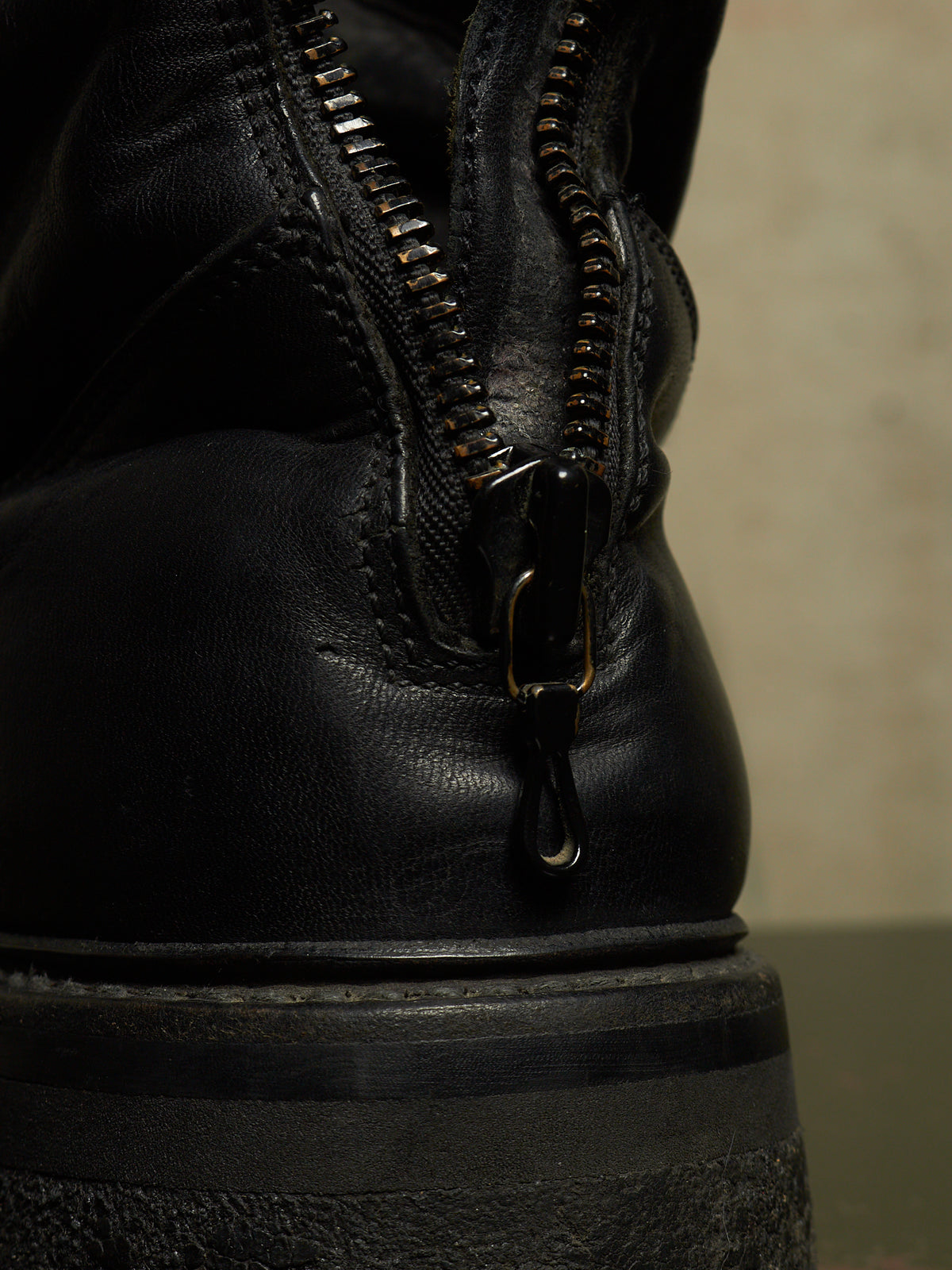 Close-up of a black leather boot showing the back zipper with metal teeth and pull tab, set against a neutral beige background.