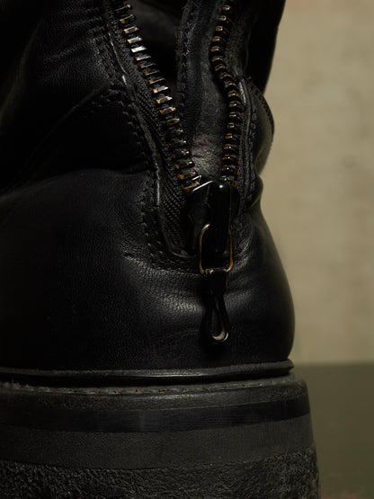 Close-up of a black leather boot showing the back zipper with metal teeth and pull tab, set against a neutral beige background.