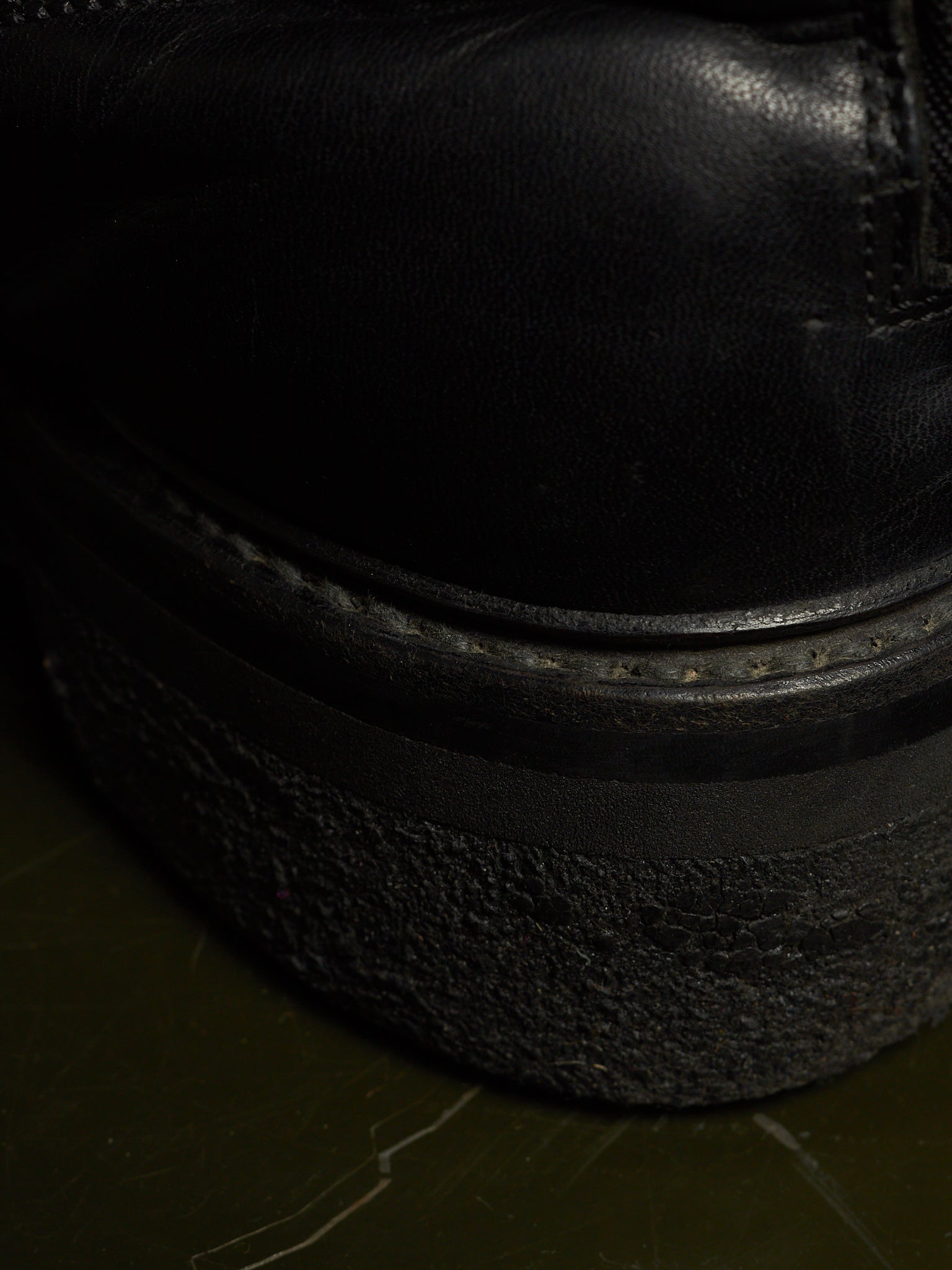 Close-up detail of a black leather boot upper meeting a thick, textured black platform sole on a dark olive-green surface.
