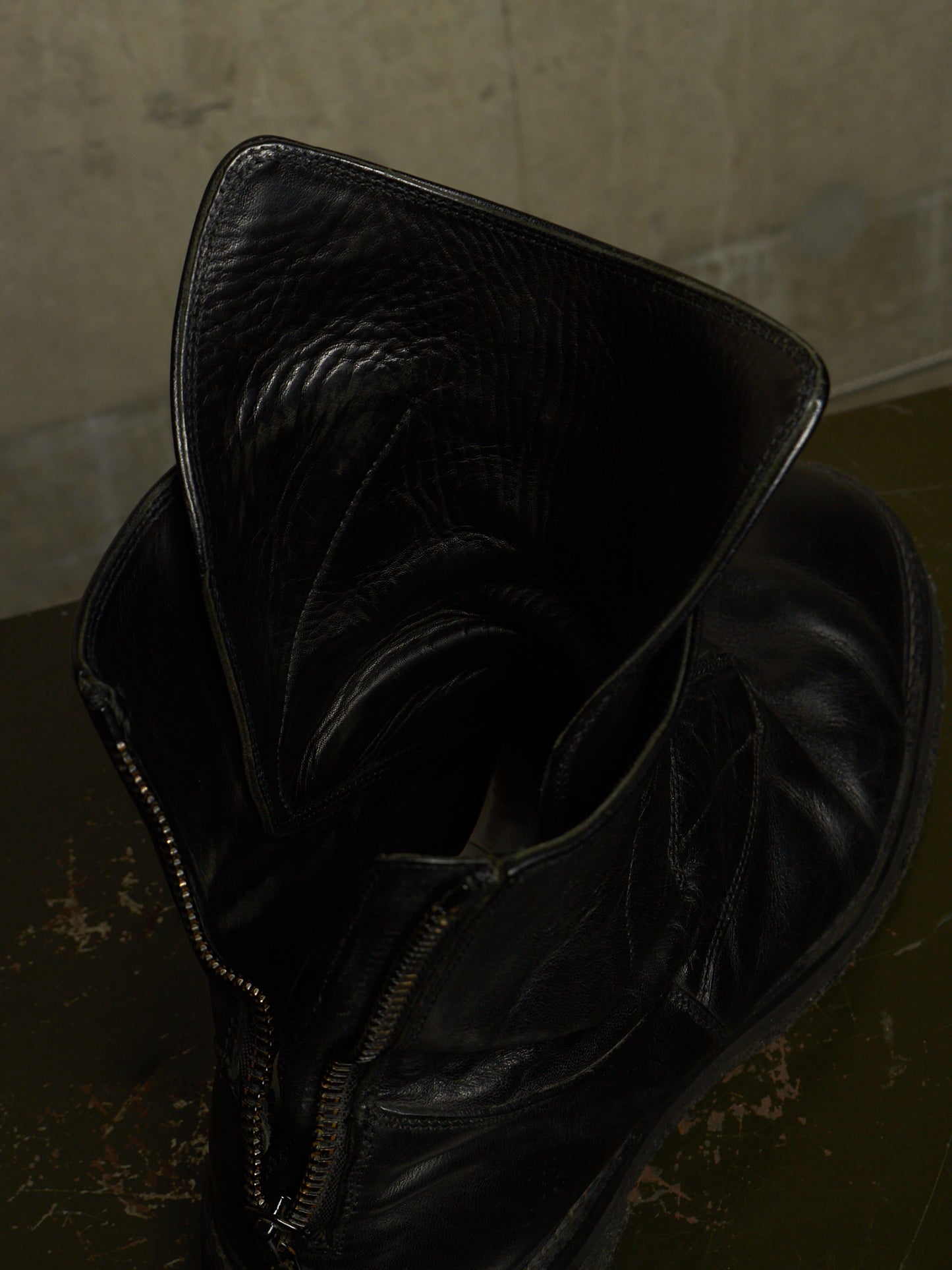 A close-up of a black leather ankle boot with a wide, folded-up collar and a side zipper, resting on a dark textured surface against a neutral concrete wall.